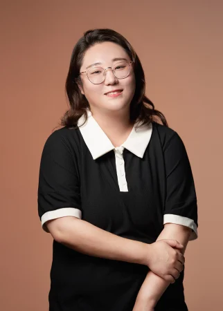 A smiling piano teacher wearing glasses and a black collared shirt with white trim, posed against a warm brown background